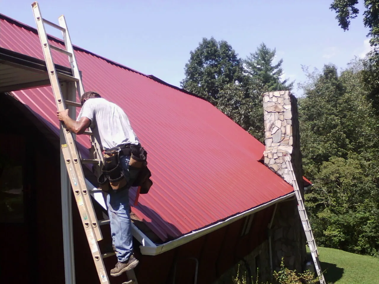 Roofer on a ladder beside a red metal roof and stone chimney — Soffit & Fascia Repair by Roofing Brothers in Pawnee